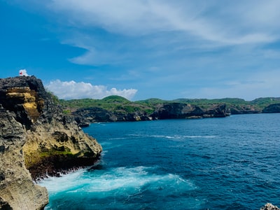 In the daytime, in the cloudy sky blue and white, blue ocean formed around the green and brown rocks
